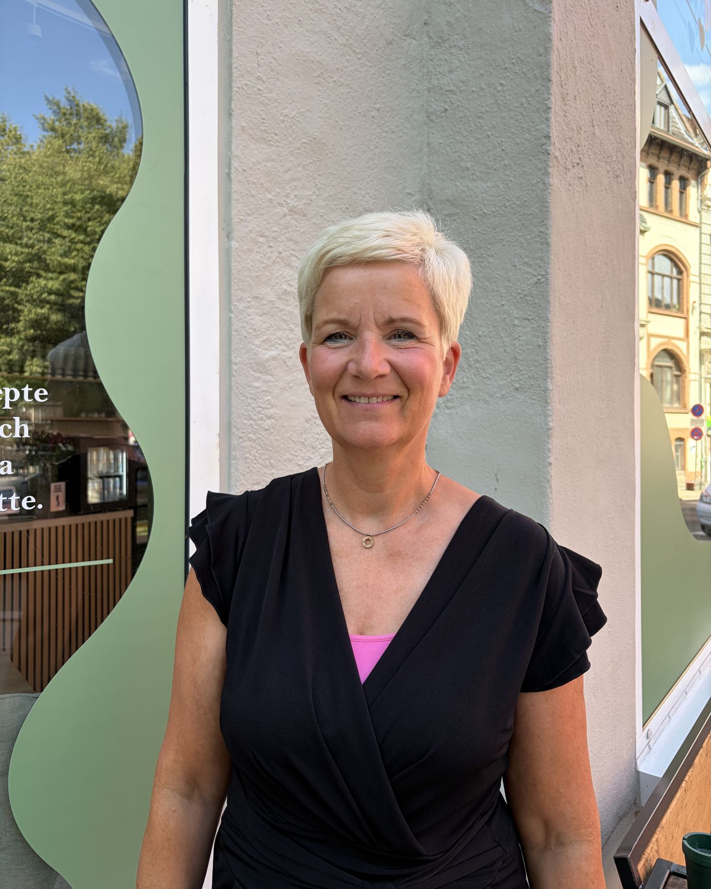 Bruni Fehse-Scholze lächelt in die Kamera – kurze helle Haare, dunkles Oberteil, vor einer hellen Hauswand neben einem grün gerahmten Schaufenster in Halle (Saale).